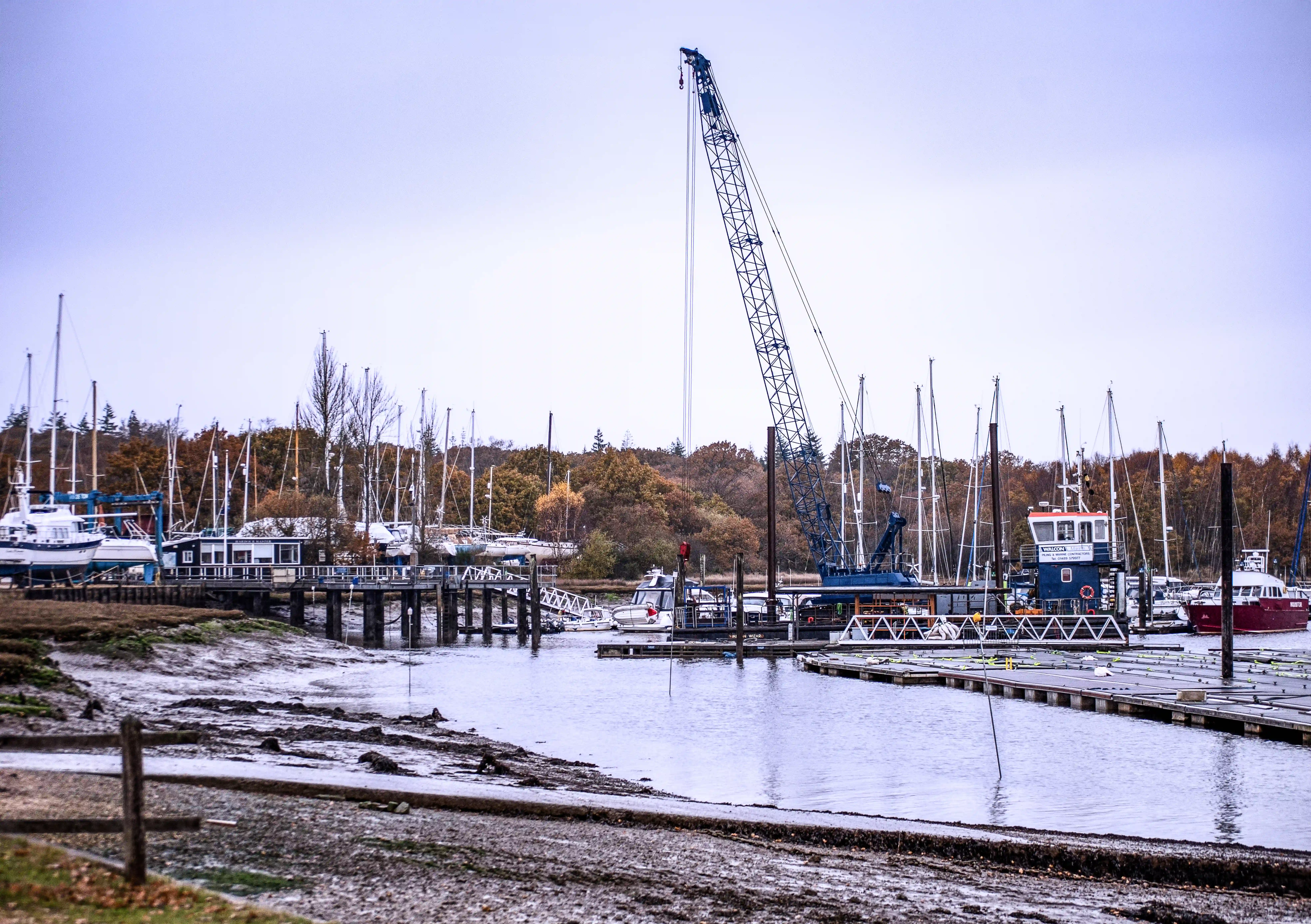 The image depicts a marina with several boats docked, including sailboats and other vessels. A large crane is prominently positioned in the center, suggesting ongoing construction or maintenance work. The marina is surrounded by a mix of trees and buildings, with a calm water body in the foreground. The overall scene suggests a quiet, possibly industrial or commercial marina setting.